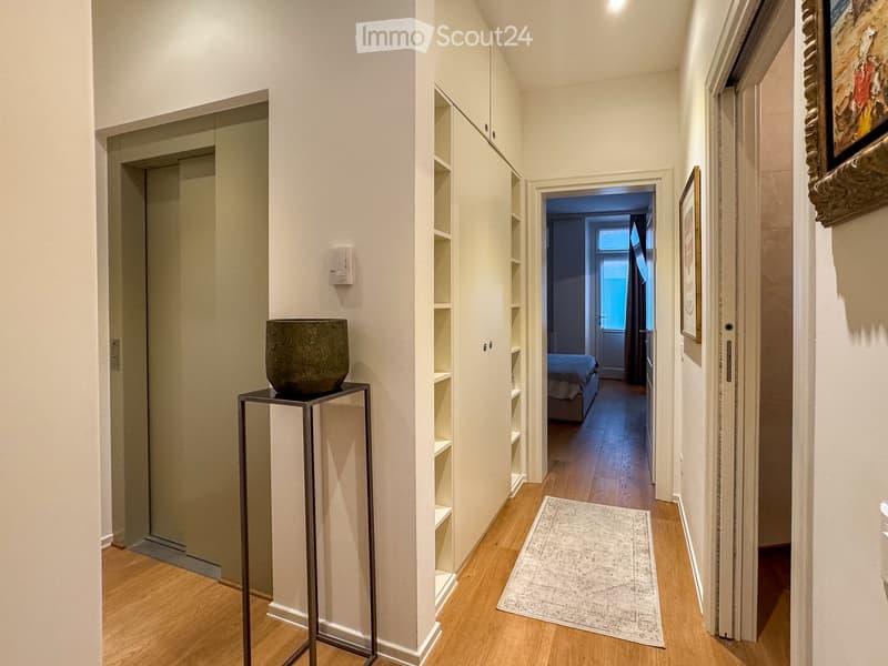 A modern hallway with light wooden flooring, white walls, built-in shelving, a small table with a decorative vase, and a glimpse of a bedroom with a bed visible at the end.