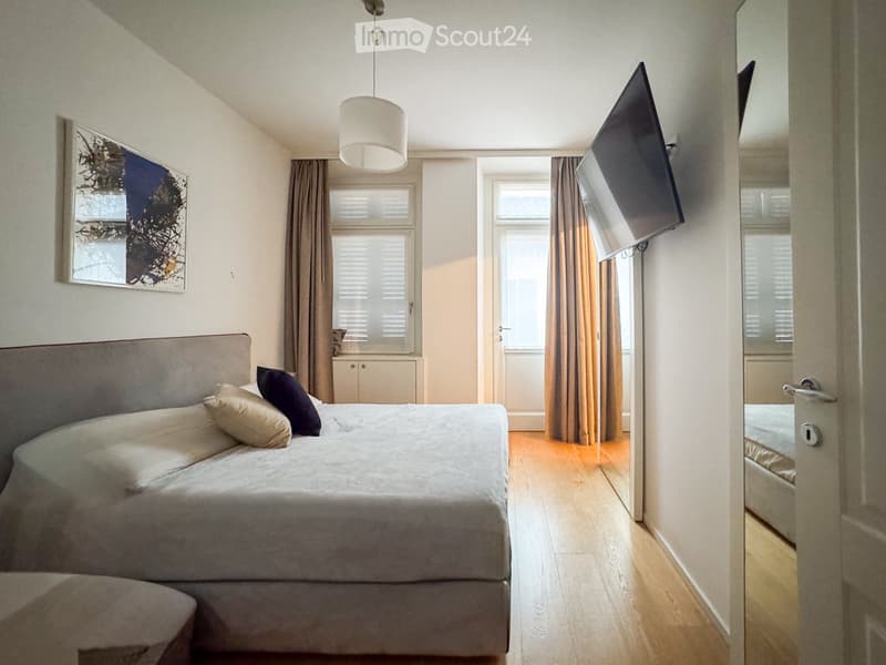 Modern bedroom with a large bed, pillows, headboard, pendant light, mirror, television, and cabinet. The room features wooden flooring and large windows with curtains.