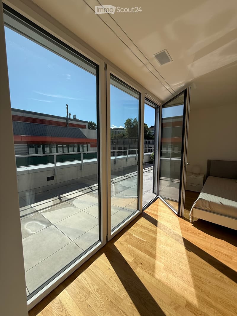 Bright bedroom with wooden flooring, large glass sliding doors leading to a balcony, and a neatly made bed.