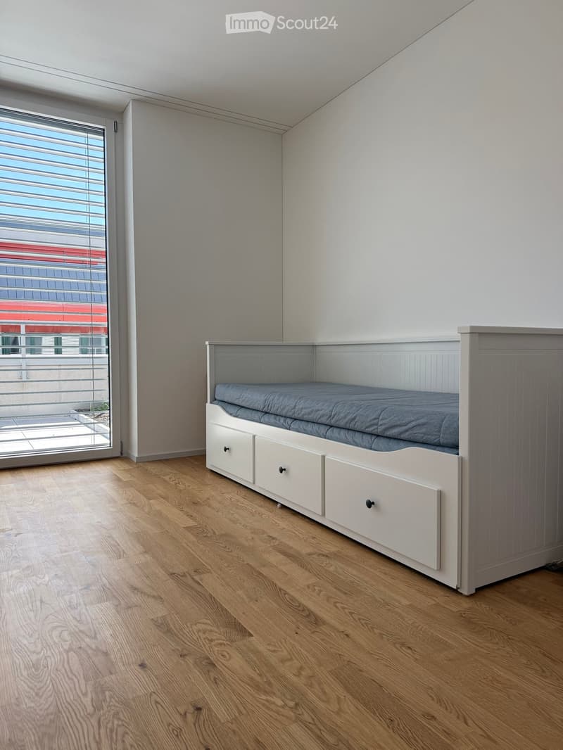 A modern bedroom with a grey bed featuring built-in drawers, light wooden flooring, and large windows with blinds.