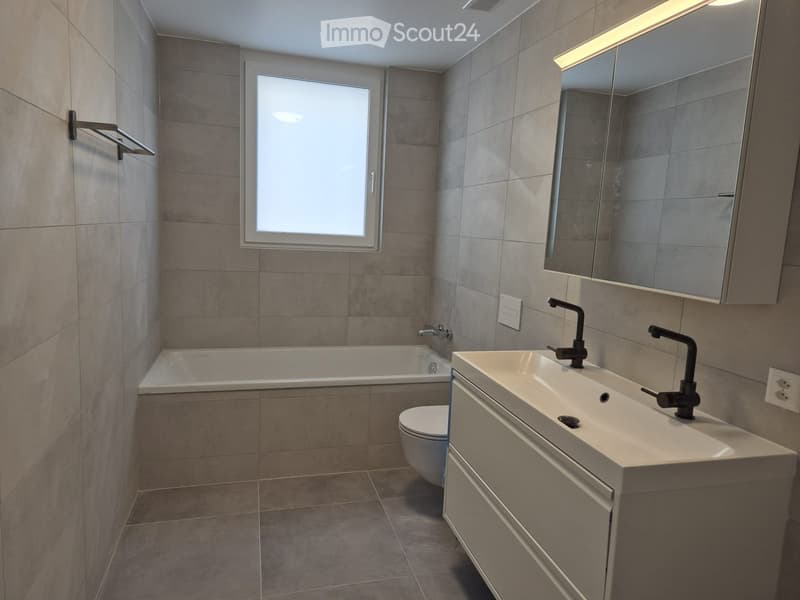 Modern bathroom with a bathtub, sink, toilet, and large mirror. Features tiled walls and floor, a window for natural light, and sleek black fixtures.
