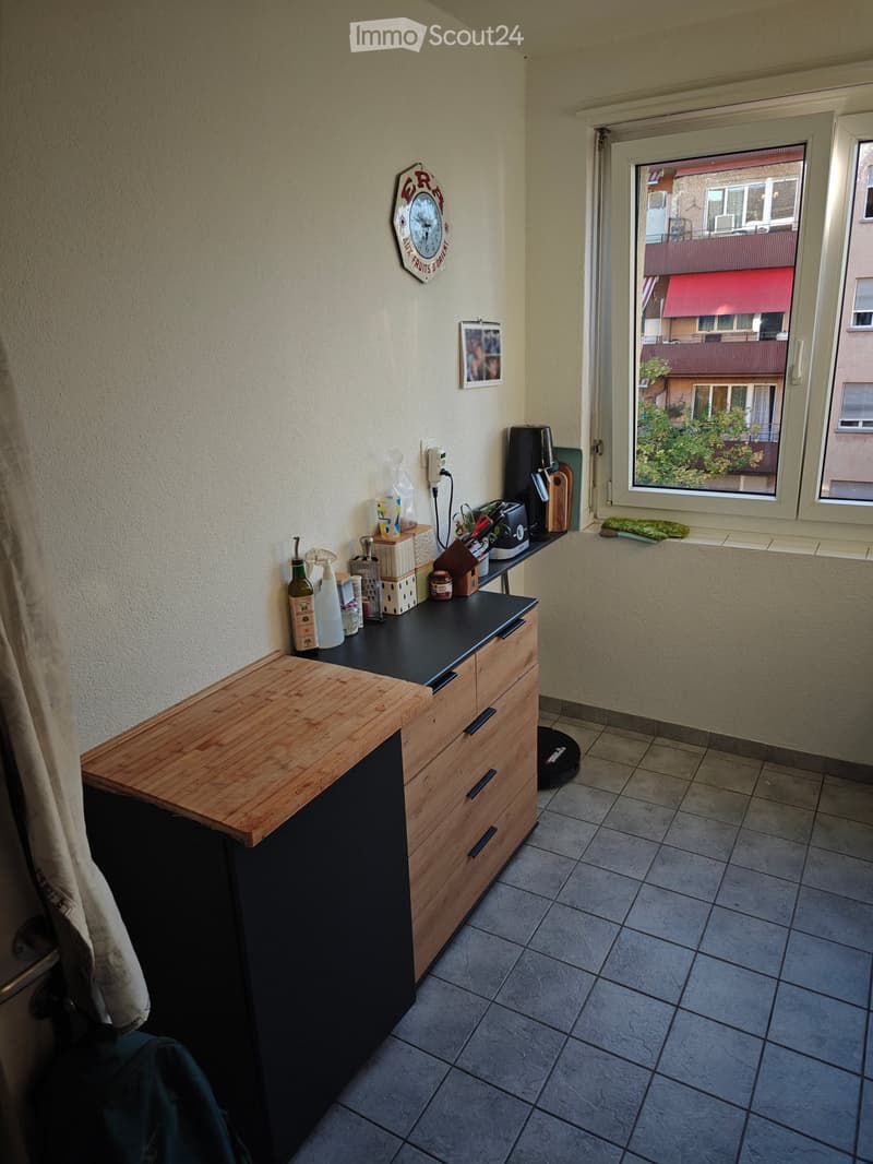 Compact kitchen with wooden counter, black countertop, drawers, and various small appliances and items on the counter. A window offers a view of the street.