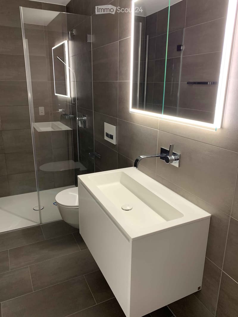 Modern bathroom with a white sink, sleek faucet, large illuminated mirror, and a glass-enclosed shower area with a toilet.