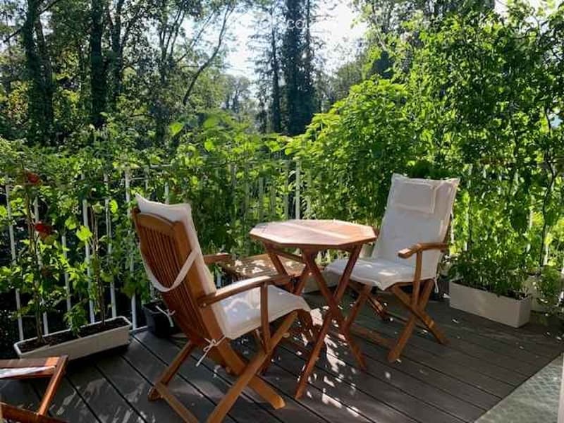 A wooden deck terrace with two white cushioned chairs and a small wooden table, surrounded by lush greenery and potted plants.