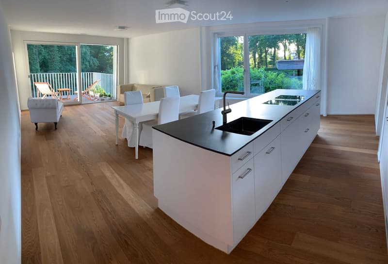 Open-plan kitchen and living area with white cabinets, a black countertop, a sink, a dining table with chairs, and a sofa. The space features wooden flooring and large windows with a view of a balcony and outdoor seating.