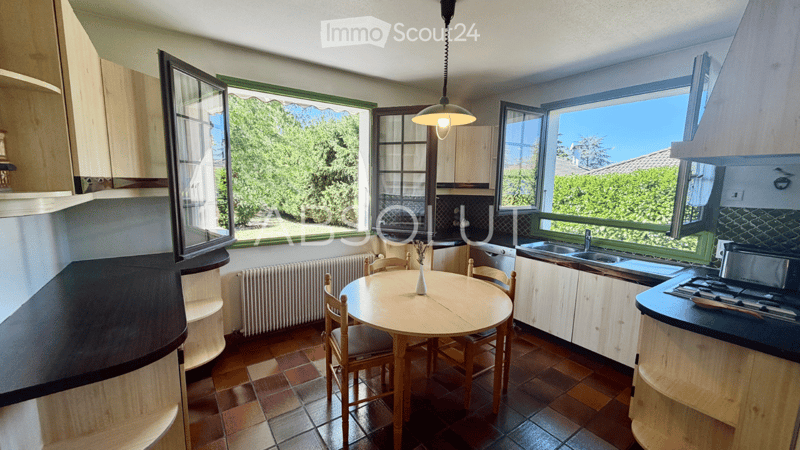 Bright kitchen with wooden cabinets, tiled floor, round dining table with chairs, sink, stove, and large windows offering a view of greenery outside.