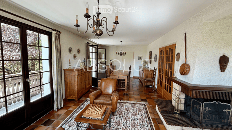 Elegant living room with a leather armchair, coffee table with chessboard, fireplace, large windows with balcony access, wooden furniture, and decorative items.
