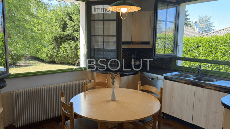 Bright kitchen with large windows offering a view of greenery, equipped with a round wooden dining table, chairs, sink, dishwasher, and light wooden cabinets.