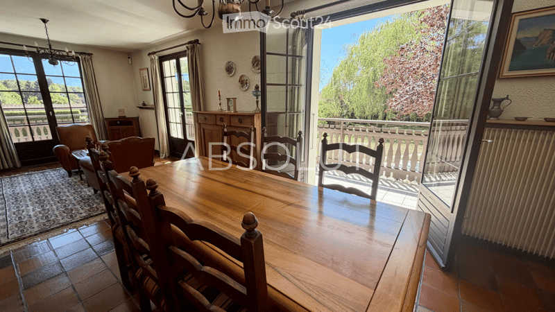 Bright dining room with large windows offering a scenic view, wooden dining table with chairs, leather sofa, armchair, cabinet, chandelier, and decorative picture frames.