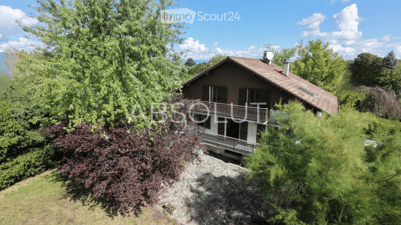 A two-story house with a brown roof and white balcony, surrounded by lush greenery and trees, offering a scenic view.