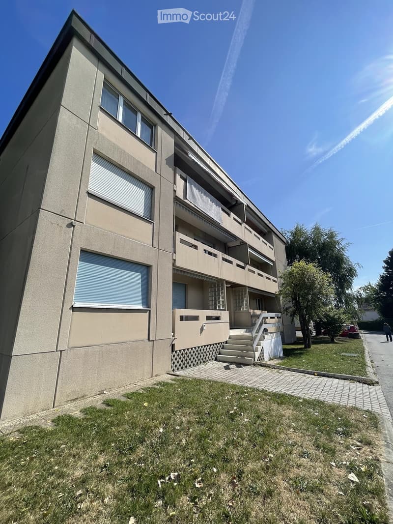 Four-story residential building with beige facade, multiple windows, balconies, and a staircase leading to the entrance.