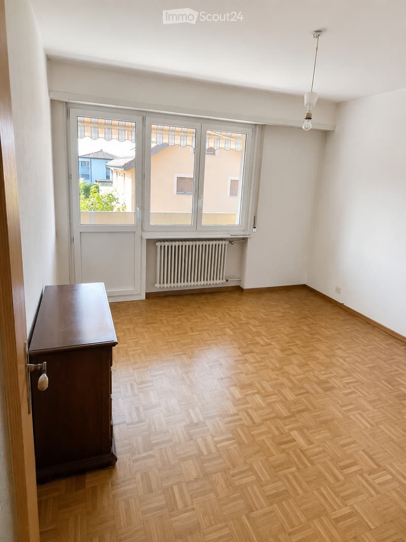 A small, unfurnished room with wooden herringbone flooring, white walls, and large windows with blinds. A single cabinet is placed against the wall near the entrance.