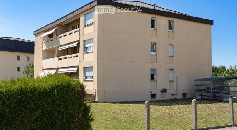 Four-story residential building with beige facade, multiple balconies, and a fenced yard with outdoor seating area.