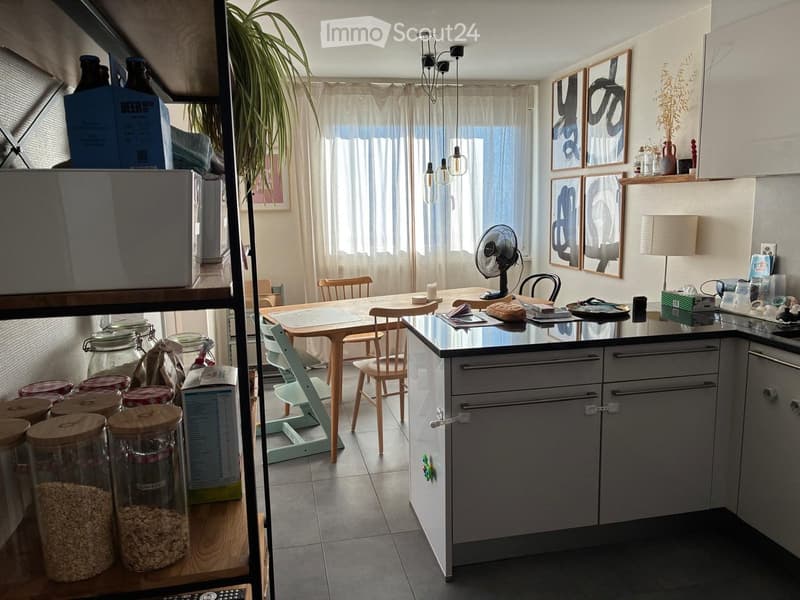 Modern kitchen with grey cabinets, a central island, a dining table with chairs, and various storage items including jars and bottles. The space features a fan, a lamp, and artwork on the walls.
