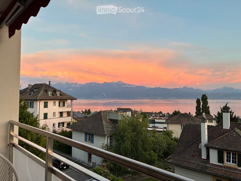 Balcony overlooking a scenic view of a lake and distant mountains at sunset, with several houses and trees in the foreground.