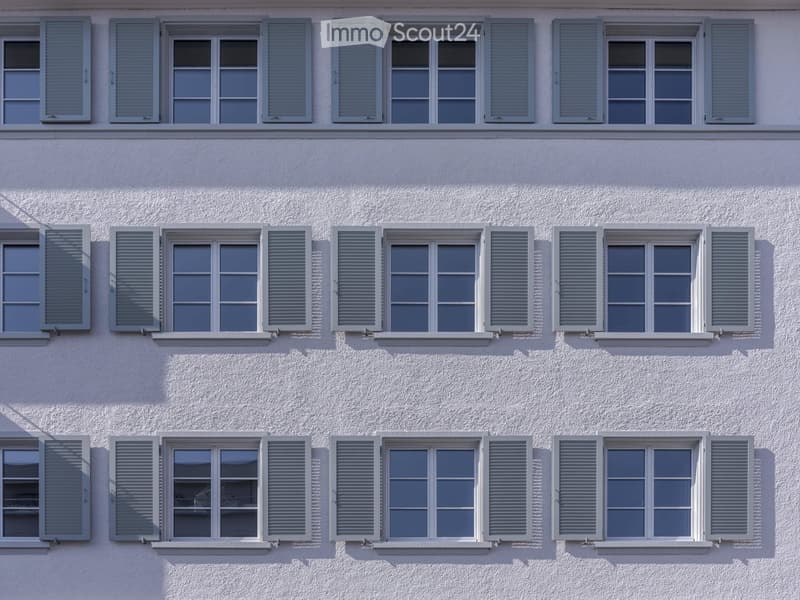 White building facade with multiple windows featuring grey shutters, arranged in a grid pattern across three visible floors.
