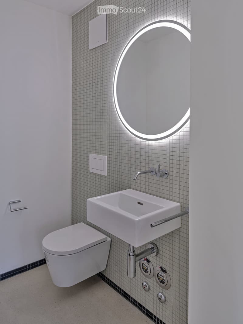 Modern bathroom with a white toilet, wall-mounted sink, chrome faucet, and illuminated round mirror.