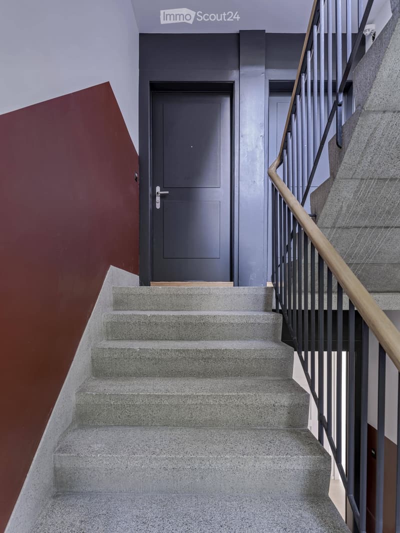 Modern staircase with concrete steps, wooden handrail, and metal balustrade, leading to a grey door.