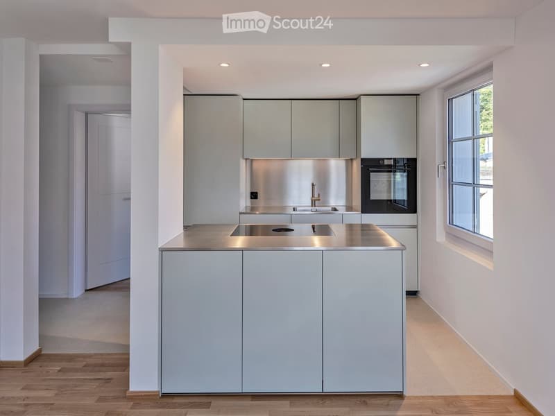 Modern kitchen with light grey cabinets, stainless steel sink, faucet, and oven. Wooden flooring and white walls.