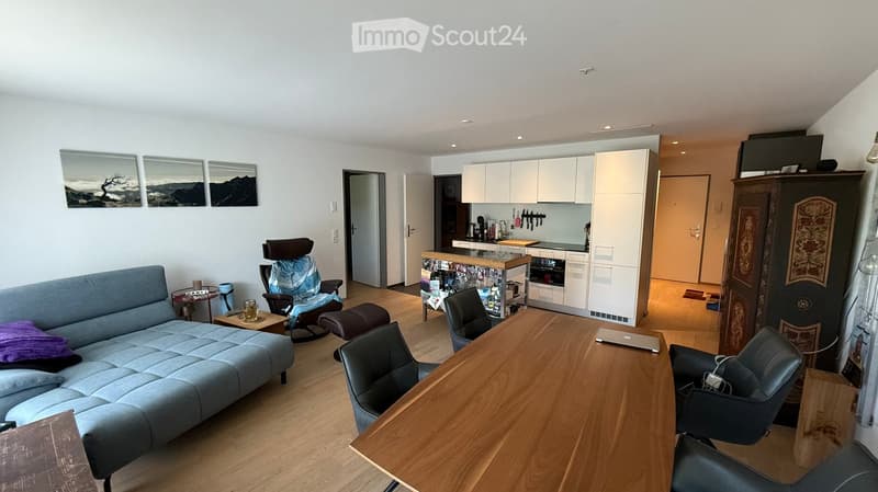 Modern living room with a blue sofa, coffee table, wooden dining table, several chairs, laptop on table, and an open kitchen area with white cabinets and appliances.