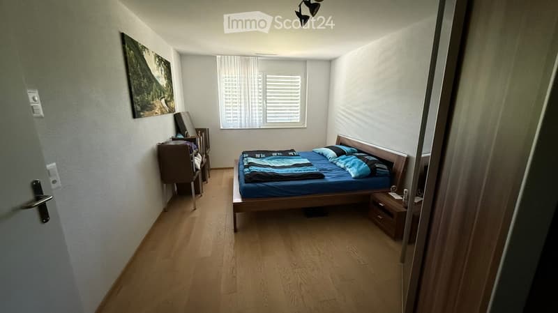 A well-lit bedroom with wooden flooring, a bed with blue and black bedding, a nightstand, a chair, and a picture frame on the wall.