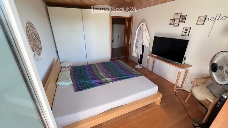 Cozy bedroom with a wooden bed, colorful striped blanket, small table with TV, wicker chair, fan, and wardrobe. Wooden flooring and white walls.