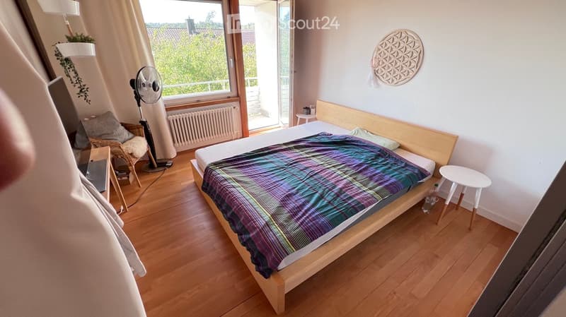 Cozy bedroom with wooden flooring, a bed with a colorful plaid blanket, a wicker chair, a standing fan, a small white stool, and a wall decoration.