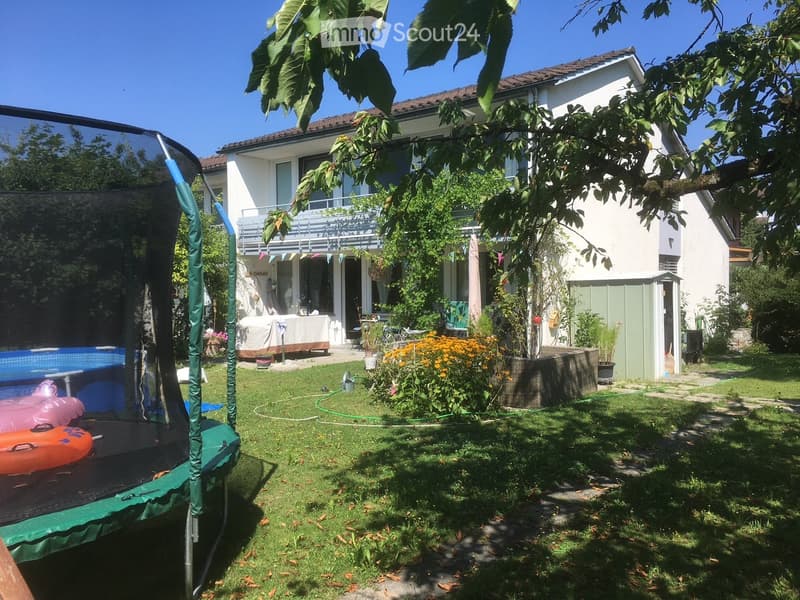Lush garden with a trampoline, swimming pool, covered patio, and colorful flowers, surrounded by a two-story house with a balcony.