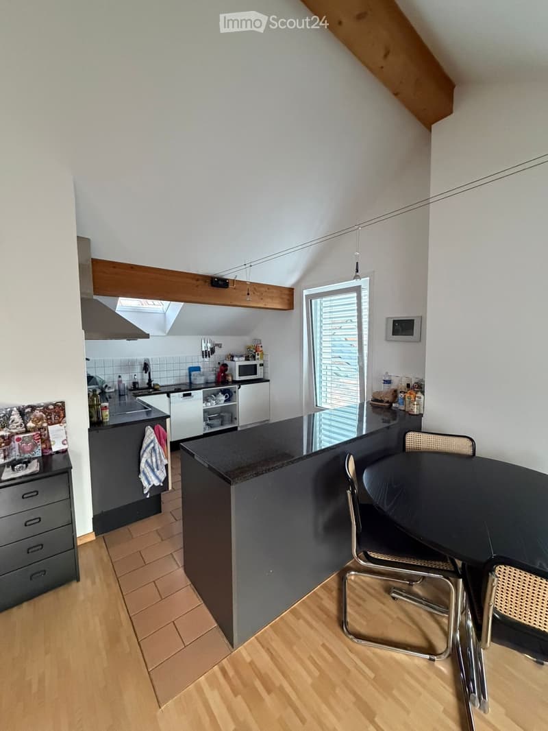 Modern kitchen with dark countertops, wooden flooring, and built-in appliances including a microwave, dishwasher, and sink. A small dining area with a table and chairs is adjacent to the kitchen island.