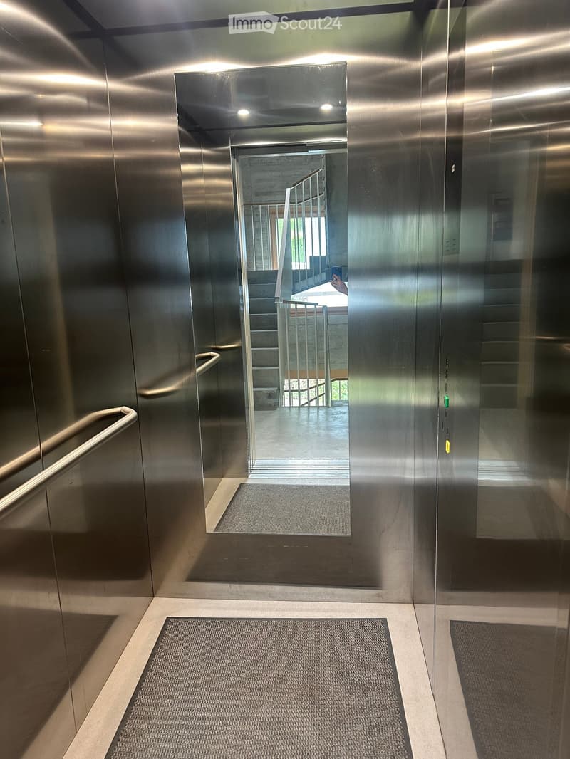 Modern hallway with metallic walls, carpeted floor, and a reflective glass door leading to a staircase area.