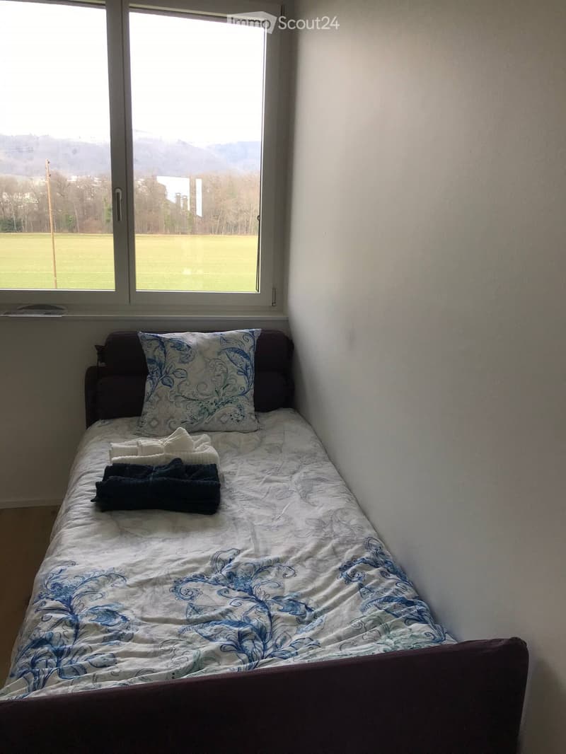 A small bedroom with a single bed, patterned bedding, and a decorative pillow, situated near a large window offering a scenic view of a grassy field and distant mountains.