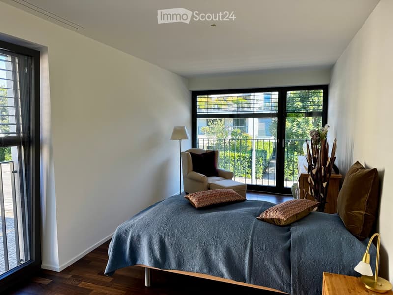 Well-lit bedroom with a large bed, pillows, a nightstand with a lamp, a chair, and a decorative vase near the balcony.