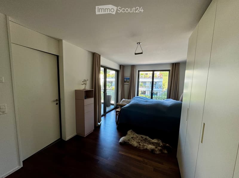 Modern bedroom with dark wooden flooring, large windows with curtains, a bed with blue bedding, a nightstand, a furry rug, and a wardrobe. A small table and chair are near the balcony door.