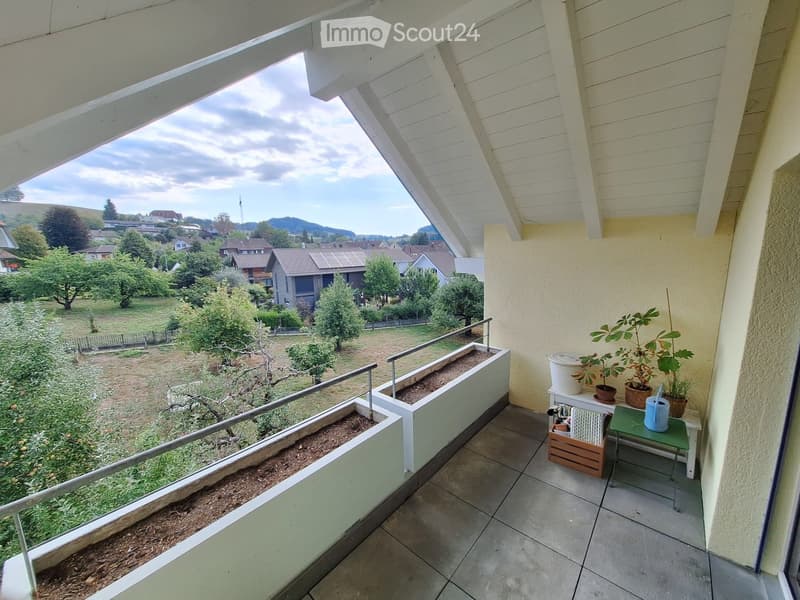 Balcony with tiled floor, metal railing, potted plants, small table, and a view of a residential area with houses and trees.