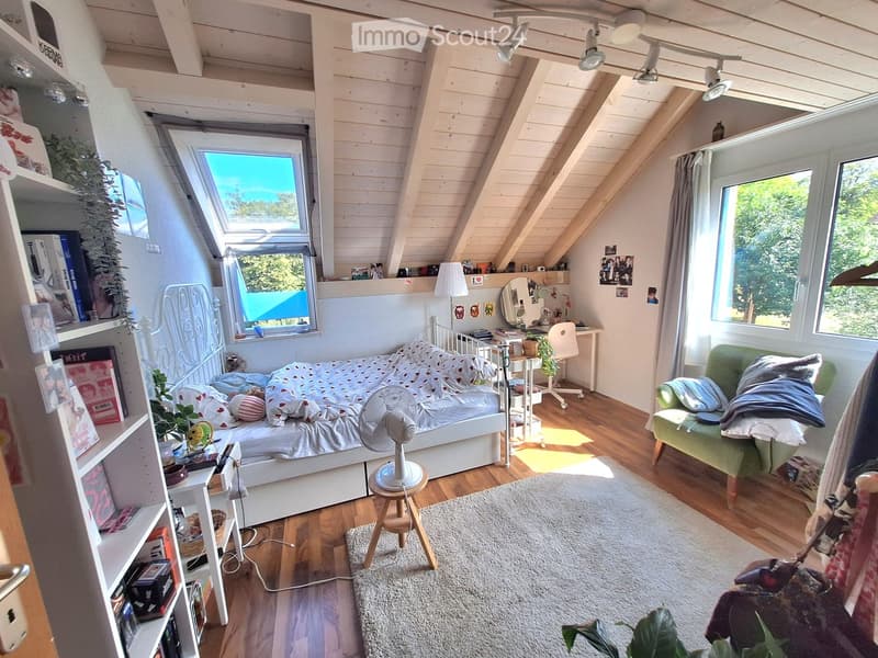 Cozy attic bedroom with wooden floors, slanted ceilings, large windows, a bed with a white metal frame, a green sofa, a small table with a fan, shelves with books and decor, and a desk area with a chair and mirror.