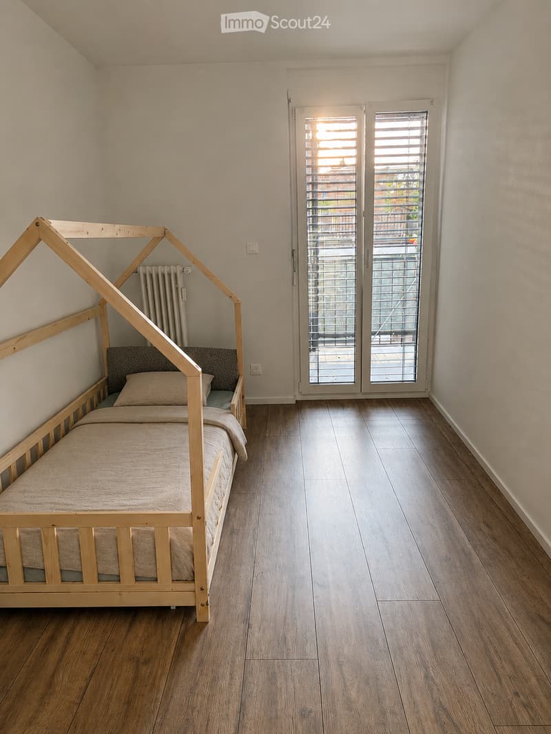 A small bedroom with a wooden bed, a radiator, and a large window with blinds leading to a balcony.