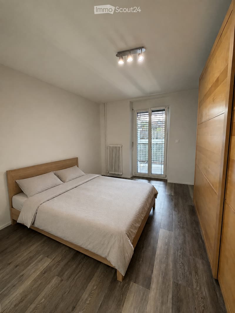 Modern bedroom with wooden flooring, a double bed with light-colored bedding, a built-in wardrobe, and a radiator near the window.