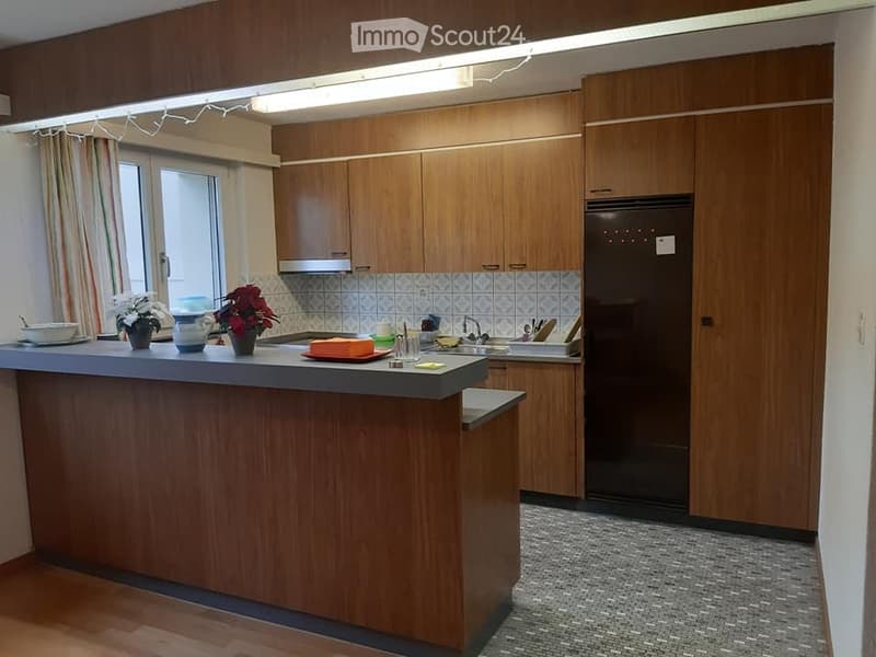 A modern kitchen with wooden cabinets, a central island, and various appliances. The island features a countertop with a bowl, vase, and flower. A refrigerator is visible on the right side, and a sink with a faucet is present in the background.
