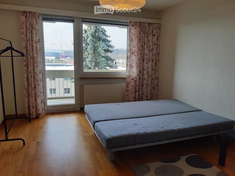 A small, furnished bedroom with a twin-sized bed, wooden flooring, large windows with curtains, and a radiator beneath the windows. A wardrobe is visible on the left side, and a patterned rug is placed near the bed.
