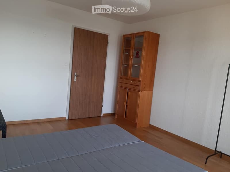 A small, unfurnished room with wooden flooring, a wooden door, and a wooden cabinet with glass doors.