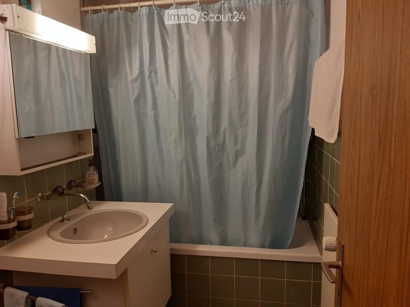 A small bathroom with a sink, mirror, and a shower curtain. The sink has a faucet and a cabinet underneath. There is a toothbrush and toothpaste on the sink counter, and a towel hanging on a rack. A bathrobe is also visible.