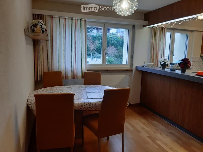 A cozy dining area with a round table and four chairs, a window with curtains, and a small kitchen counter with a few items on it.