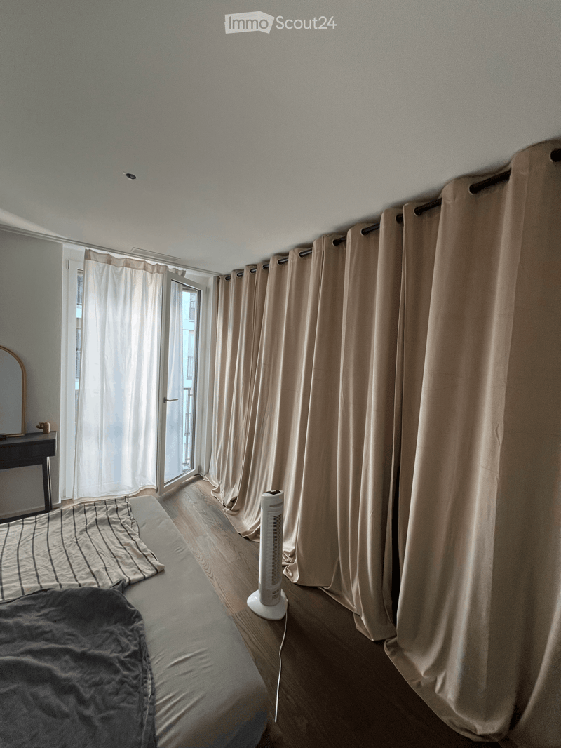 A modern bedroom with wooden flooring, large windows with beige curtains, a bed with grey bedding, a fan, and a small table with a mirror.