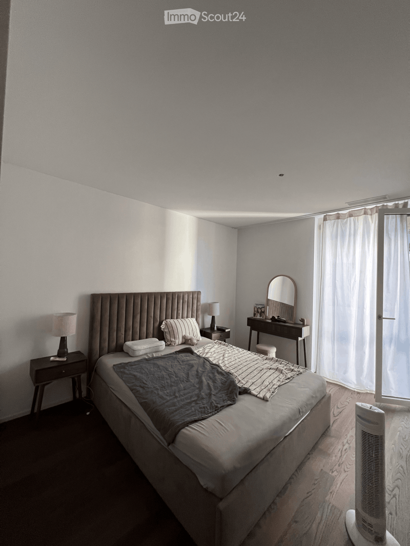 A modern bedroom with a large bed, nightstands, lamps, a mirror, and a fan. The room features light-colored walls and wooden flooring.