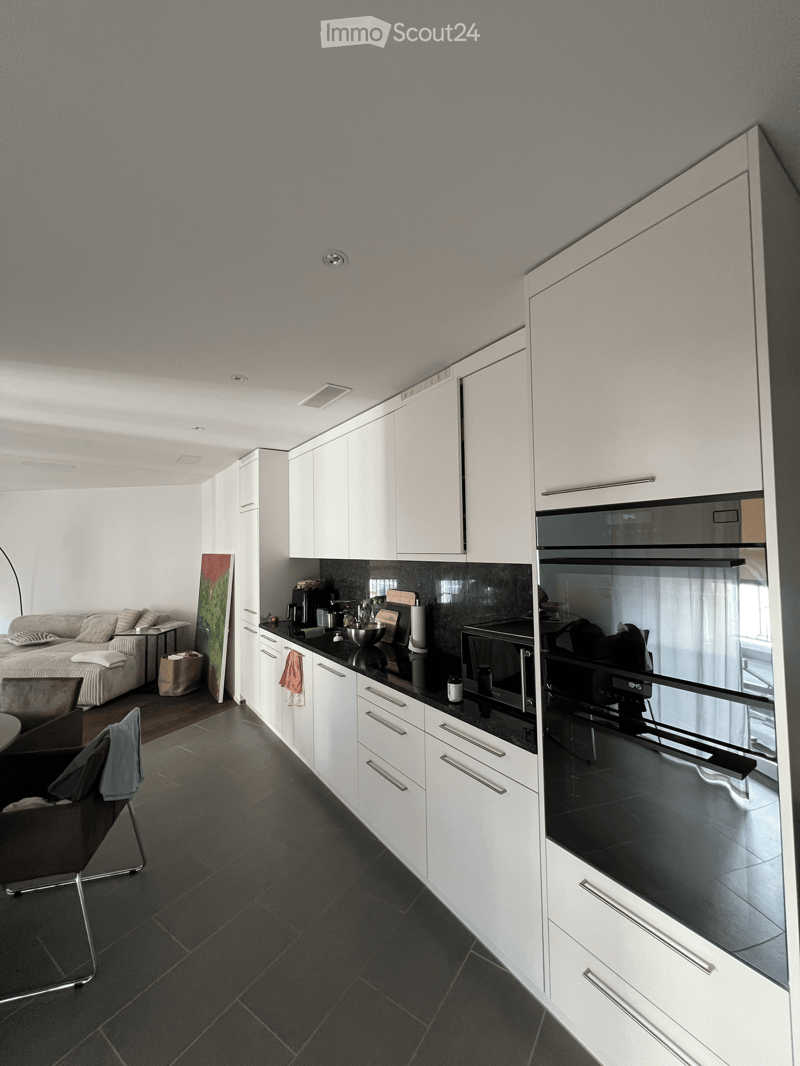 Modern kitchen with white cabinets, black countertops, and stainless steel appliances. A sofa and chair are visible in the adjacent living area.