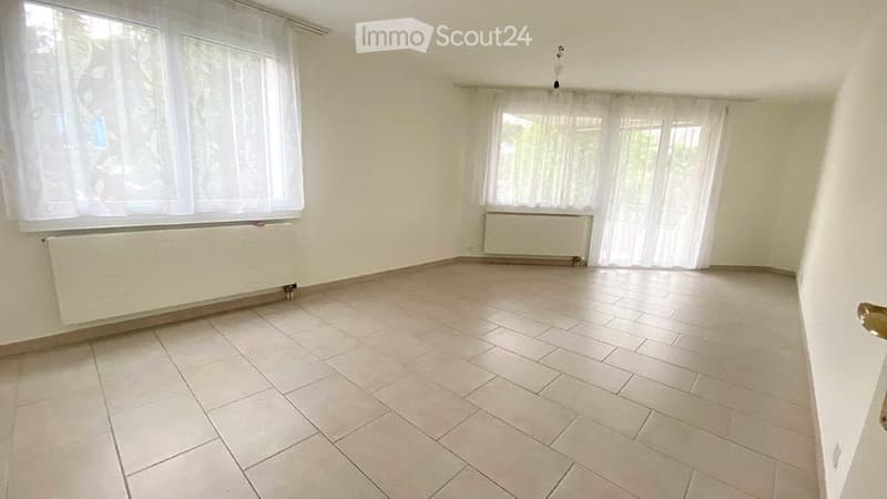 Empty room with tiled floor, white walls, large windows with white curtains, and radiators under the windows.