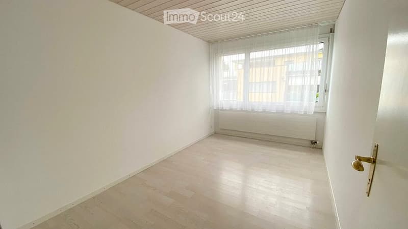 Empty room with white walls, light wooden flooring, and a window with sheer curtains. No furniture or equipment present.