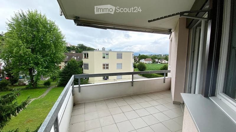 Balcony with tiled floor, metal railing, and a view of a green lawn, trees, and neighboring buildings.