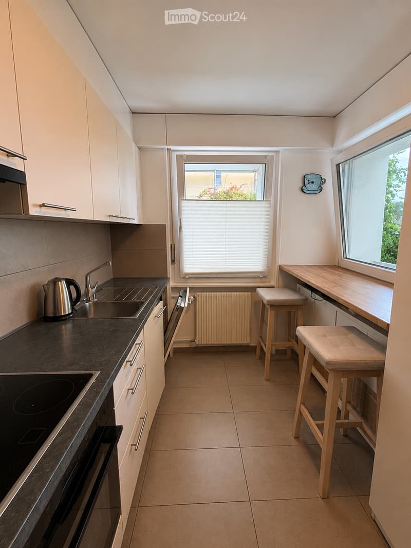 Modern kitchen with white cabinets, dark countertops, stainless steel appliances, and a small dining area with two stools and a table.