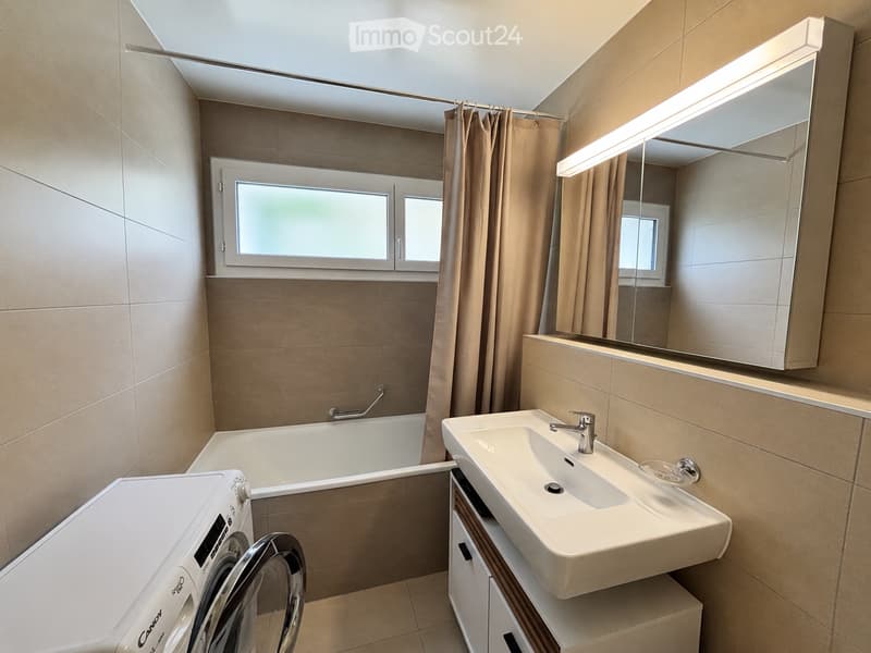 Modern bathroom with beige tiles, a bathtub, a sink with a mirror, a washing machine, and a window with a curtain.